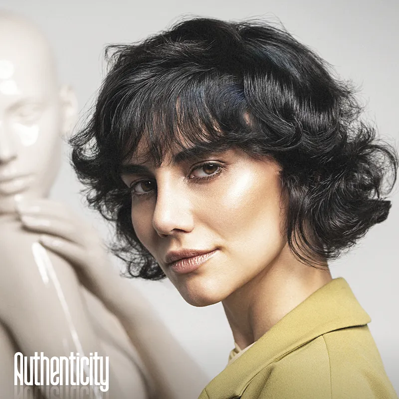 Donna con capelli neri corti e ricci, trucco naturale e giacca verde chiaro, sfondo neutro con statua bianca e scritta Authenticity.