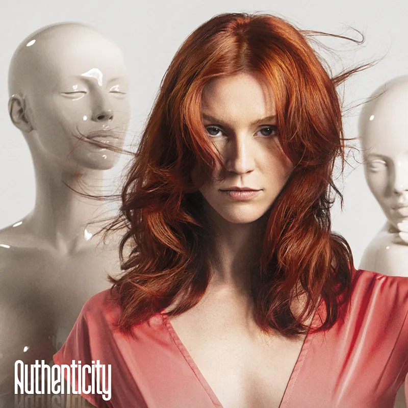 Donna con capelli rossi mossi e abito rosa davanti a due manichini bianchi, con il logo Authenticity in basso a sinistra